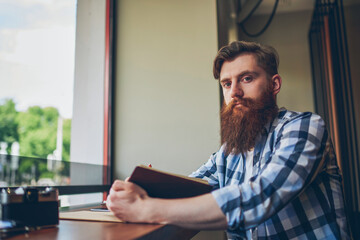 Fototapeta premium Half length portrait of skilled bearded student with textbook in hand looking at camera while sitting in coffee shop and preparing for upcoming exams.Publicity area for your advertising text message