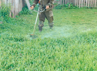 Asian man in a mosquito suit with a professional brush cutter trimmer mows the grass in the yard....