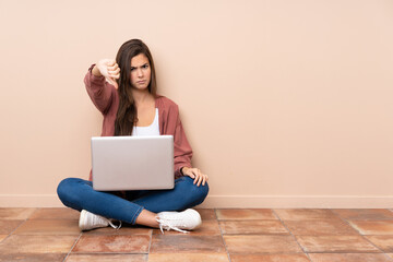 Teenager student girl sitting on the floor with a laptop showing thumb down with negative expression