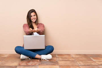 Teenager student girl sitting on the floor with a laptop holding copyspace imaginary on the palm to insert an ad