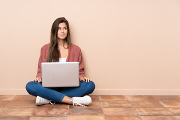 Teenager student girl sitting on the floor with a laptop making doubts gesture looking side