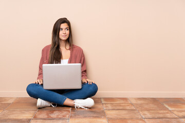 Naklejka premium Teenager student girl sitting on the floor with a laptop making doubts gesture looking side