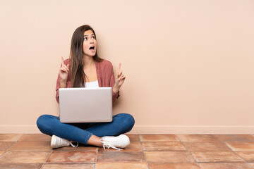Teenager student girl sitting on the floor with a laptop surprised and pointing up - obrazy, fototapety, plakaty