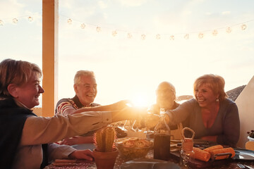 Happy senior friends dining and drinking red wine on house terrace - Mature people having fun cheering together at dinner party on sunset time - Food and elderly friendship lifestyle concept