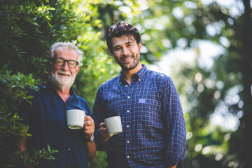 An adult hipster son and senior father drinking coffee in the garden at home, happy talking in the morning