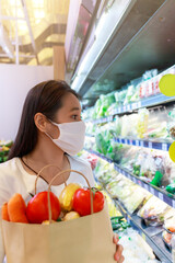 Asian woman wearing protective face mask hold paper shopping bag with fruits and vegetables in supermarket . Girl, looking grocery to buy  some fruit. New normal after covid-19. Family concept.