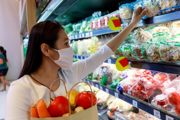 Asian woman wearing protective face mask hold paper shopping bag with fruits and vegetables in supermarket . Girl, looking grocery to buy  some fruit. New normal after covid-19. Family concept.