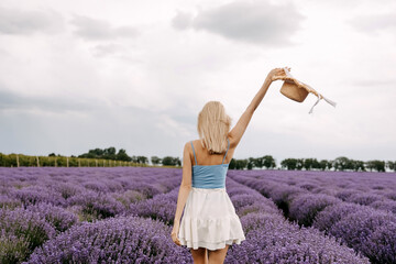 Young romantic woman standing in lavender field, holding a straw hat up in the air.