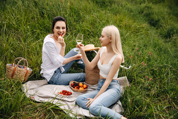 Two young women sitting on a blanket at a picnic, chatting, drinking white wine and eating fruits, outdoors in a field.