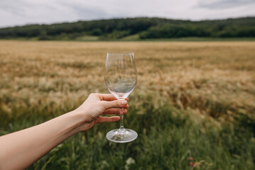 Female hand holding an empty glass of wine on wheat field background in mountains.