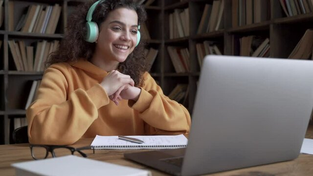 Hispanic teen girl school pupil college student wear headphones learning online with teacher tutor waving hand using video conference call by webcam on laptop computer. Videoconference education.