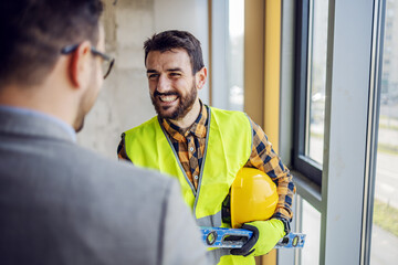 Smiling bearded contractor talking to businessman while standing in building in construction...