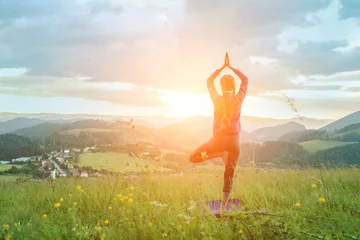 Rolgordijnen Yoga in nature, fresh air in park. Sporty girl practicing pose, stretching health on top of mountain in meadow at sunrise, zen wellness. Teamwork, good mood and healty life. © Andrii IURLOV