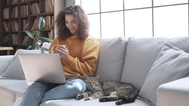 Happy latin teen girl looking at laptop laugh relax with cat on couch at home. Smiling young hispanic woman watching movie holding computer having fun with pet drinking tea sit on sofa in living room.