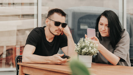 A young woman and man sits on an open veranda in a cafe and uses a smartphone while waiting for order and takeaway. 