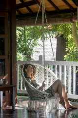 Woman sitting in the wicked hanging chair with pillow at the balcony with green nature tropical...