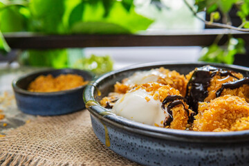 Fried white ice cream on blue plate, grey table with flowers near window. Natural day light in the kitchen. Homemade dessert. Copy space. Hot ice in corn breading with caramel and chocolate sauce.