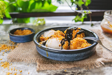 Fried white ice cream on blue plate, grey table with flowers near window. Natural day light in the kitchen. Homemade dessert. Copy space. Hot ice in corn breading with caramel and chocolate sauce.