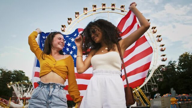Mixed raced females in sunglasses are smiling and dancing, holding flag of USA and waving it while standing in park against a ferris wheel. Slow motion