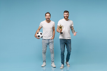 Excited two young men guys friends in white t-shirt posing isolated on pastel blue background in studio. Sport leisure lifestyle concept. Cheer up support favorite team with soccer ball, football cup.