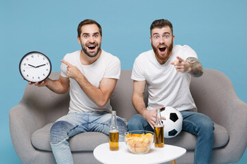 Excited men guys friends in white t-shirt sit on couch isolated on pastel blue background. Sport leisure concept. Cheer up support favorite team with soccer ball poin index fingers on clock, camera.