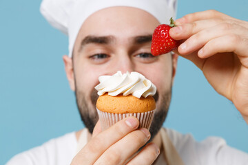 Cropped image of young bearded male chef or cook baker man in apron white t-shirt toque chefs hat isolated on blue background studio. Cooking food concept. Decorating dessert cupcake with strawberry.