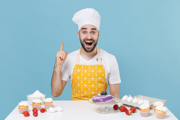 Excited young male chef or cook baker man in apron white t-shirt toque chefs hat cooking at table isolated on blue background studio. Cooking food concept. Holding index finger up with great new idea.