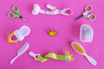 Flat lay on baby care items - scissors, hairbrushes, pacifiers, pacifier holders, nasal aspirator and nibbler- on pink background with little yellow crown for little princess on Birthday.