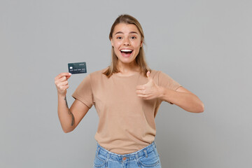 Excited young blonde woman girl in casual beige t-shirt posing isolated on gray wall background studio portrait. People lifestyle concept. Mock up copy space. Hold credit bank card showing thumb up.