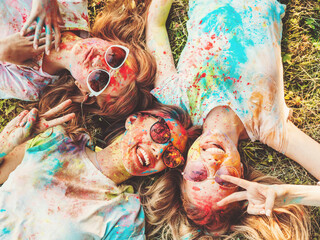 Three happy beautiful girls making party at Holi colors festival in summer time.Young  smiling women friends having fun after music event at sunset.Models lying on the grass in sunglasses.