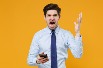 Angry young business man in classic blue shirt tie isolated on yellow background. Achievement career wealth business concept. Mock up copy space. Using mobile phone spreading hands screaming swearing.