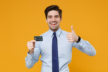 Cheerful young business man in classic blue shirt tie posing isolated on yellow background. Achievement career wealth business concept. Mock up copy space. Hold credit bank card, showing thumb up.