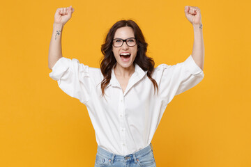Happy young brunette business woman in white shirt glasses isolated on yellow background. Achievement career wealth business concept. Mock up copy space. Clenching fists like winner, rising hands.