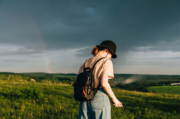 Stylish hiker girl in casual clothes and backpack walks on the mountain on a background of rainbow and blue rain clouds. Domestic tourism.