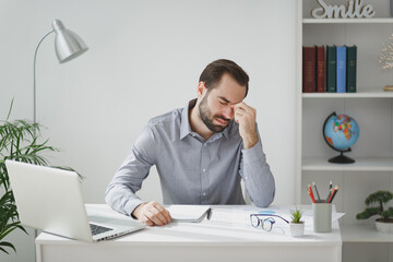 Exhausted tired young bearded business man in gray shirt sitting at desk work on laptop pc computer in light office on white wall background. Achievement business career concept. Put hand on nose.