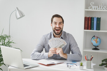 Excited business man in gray shirt sit at desk work on laptop pc computer in light office on white wall background. Achievement business career concept. Hold fan of cash money in dollar banknotes.