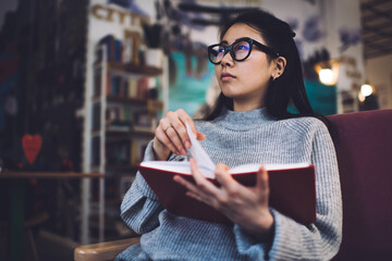 Serious chinese woman in casual wear and spectacles for vision correction turning pages on interesting book, pensive smart female 20s asian student spending time on education in comfortable interior