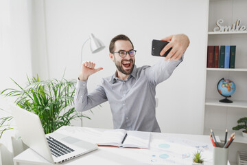 Naklejka premium Excited business man in gray shirt sit at desk work on laptop in light office on white wall background. Achievement business career concept. Doing selfie shot on mobile phone, point thumb on himself.