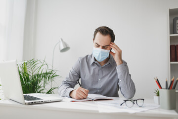 Young business man in gray shirt sterile face mask sit at desk work on laptop in light office on white wall background. Achievement business career concept. Writing notes in notebook put hand on head.
