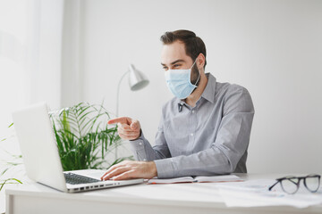 Young business man in gray shirt sterile face mask sit at desk in office on white wall background. Achievement business career concept. Work pointing finger on laptop pc computer making video call.