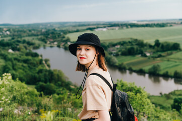 Positive girl tourist in casual clothes standing on a hill against the backdrop of the lake and looking at the camera with a smile on his face. Smiling hipster girl is engaged in domestic tourism