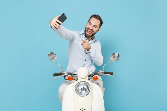 Funny young bearded man guy in casual light shirt driving moped isolated on pastel blue background. Driving motorbike transportation concept. Doing selfie shot pointing index finger on mobile phone.