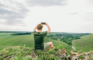 Naklejka na ściany i meble Young male hiker sits on top of a mountain and takes a photo of beautiful landscape on smartphone. Young man sits on a mountain rock and takes a photo of the Ukrainian landscape. Travel to Ukraine.