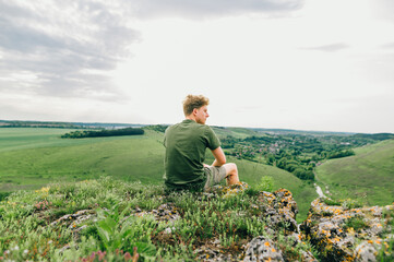 Naklejka na ściany i meble A young man in casual clothes sits on a cliff on a rock and looks at the beautiful Ukrainian landscape from a height. A tourist is resting on top of a mountain in Ukraine. Tovtry.