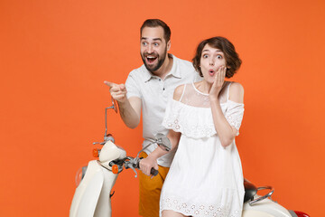 Shocked young couple two friends guy girl in white summer clothes sitting on moped isolated on orange background. Driving motorbike transportation concept. Point index finger aside, put hand on cheek.