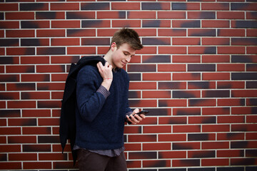 Fototapeta na wymiar Stylish young man using smartphone with pleasure on street against brown brick wall in city