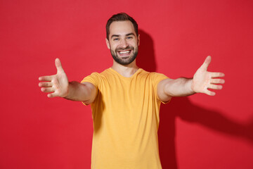 Smiling young bearded man guy in casual yellow t-shirt posing isolated on red wall background studio portrait. People lifestyle concept. Mock up copy space. Standing with outstretched hands for hugs.