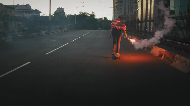 Young man in flag of USA tied on his neck is riding skateboard along deserted street with burning red signal flare in hand. Slow motion, back view