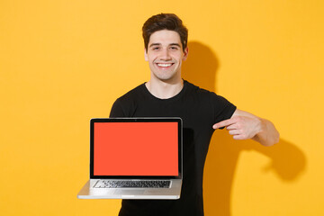 Smiling young man guy 20s in casual black t-shirt posing isolated on yellow background. People lifestyle concept. Mock up copy space. Point index finger on laptop pc computer with blank empty screen.