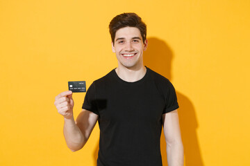 Smiling young man guy 20s in casual black t-shirt posing isolated on yellow background studio portrait. People sincere emotions lifestyle concept. Mock up copy space. Hold in hand credit bank card.